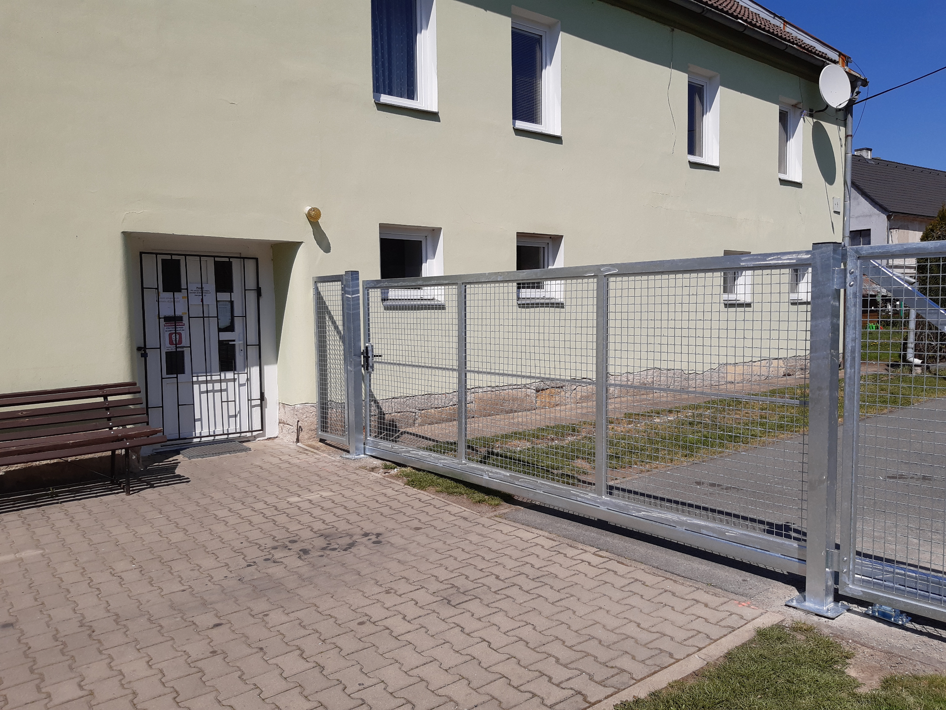 Samostatná brána — pozinkovaná brána s tahokovem vedle vstupu do budovy s lavičkou
